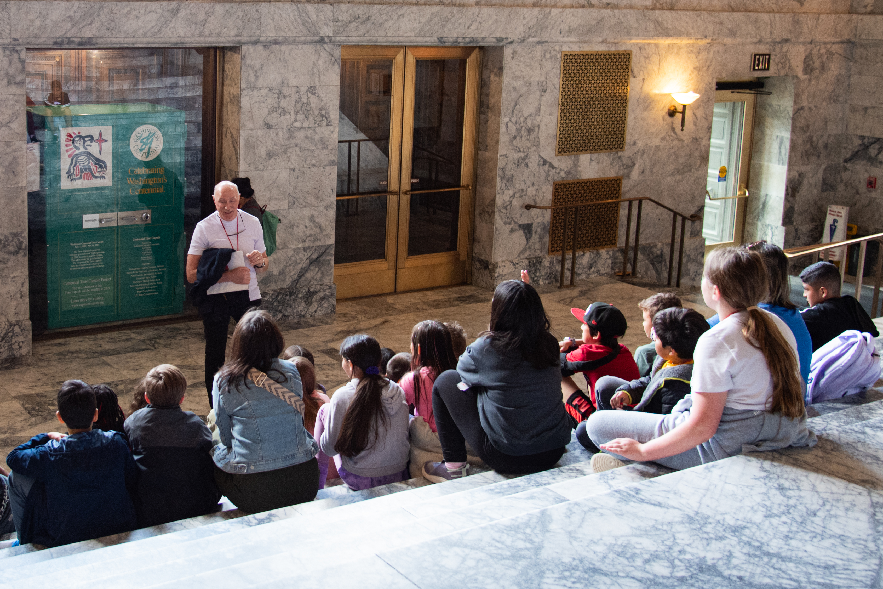 A tour guide smiling at a group of children sitting on marble steps inside the Legislative Building.