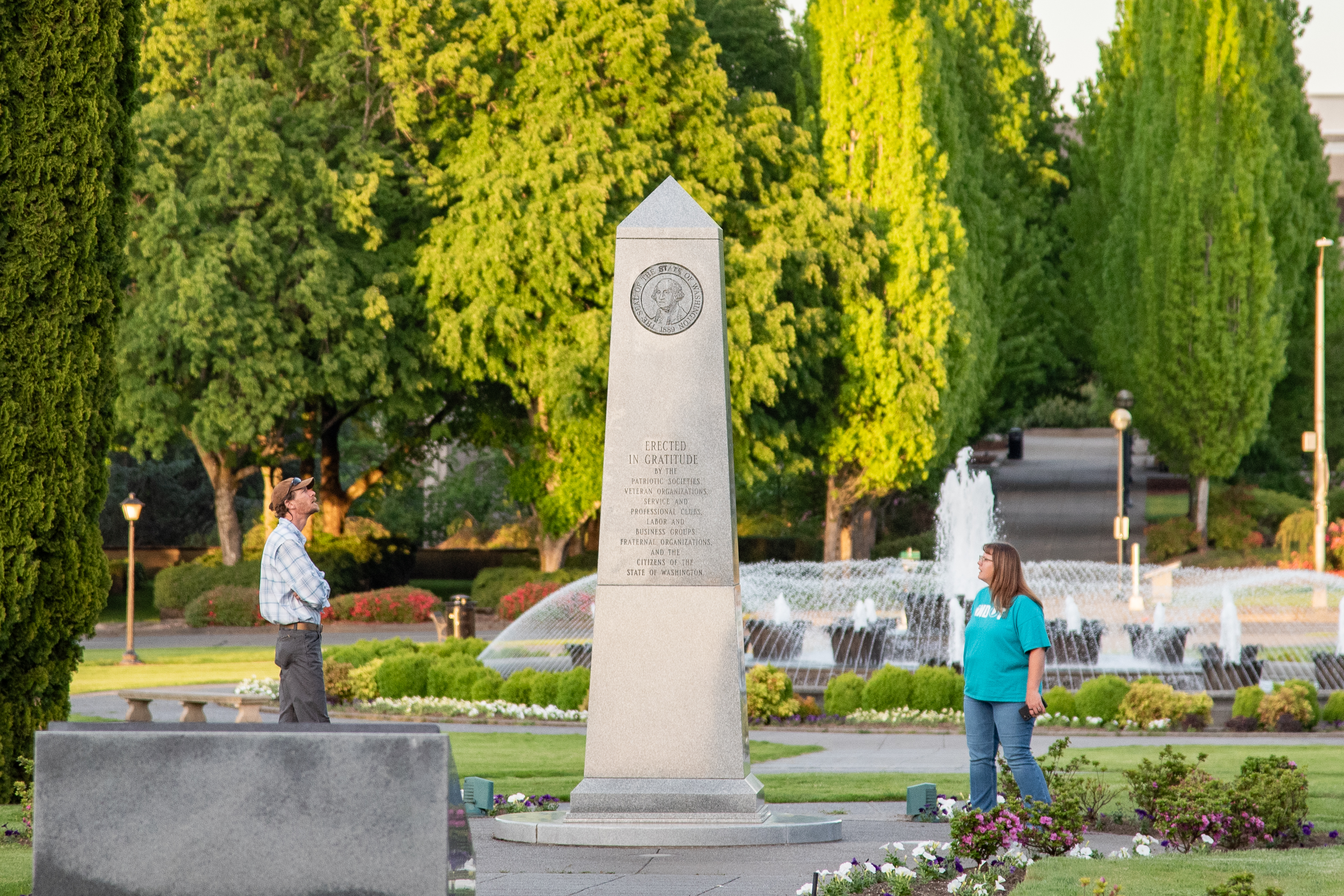 A man and a woman standing on both sides of the Medal of Honor Memorial, reading its inscriptions, with the Tivoli Fountain and green trees in the background.