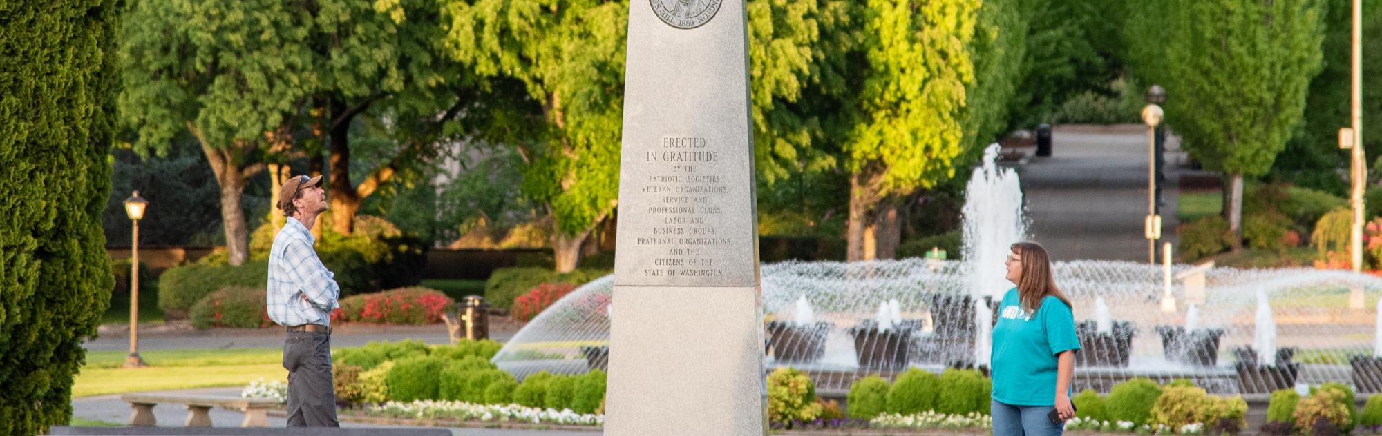 A man and a woman standing on both sides of the Medal of Honor Memorial, reading its inscriptions, with the Tivoli Fountain and green trees in the background.