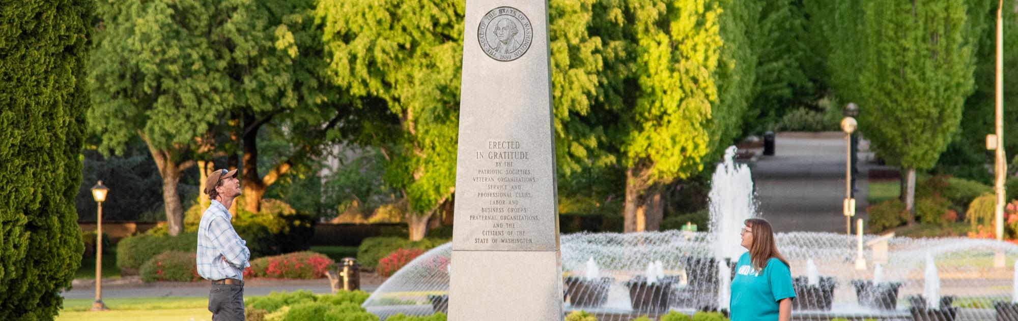 A man and a woman standing on both sides of the Medal of Honor Memorial, reading its inscriptions, with the Tivoli Fountain and green trees in the background.