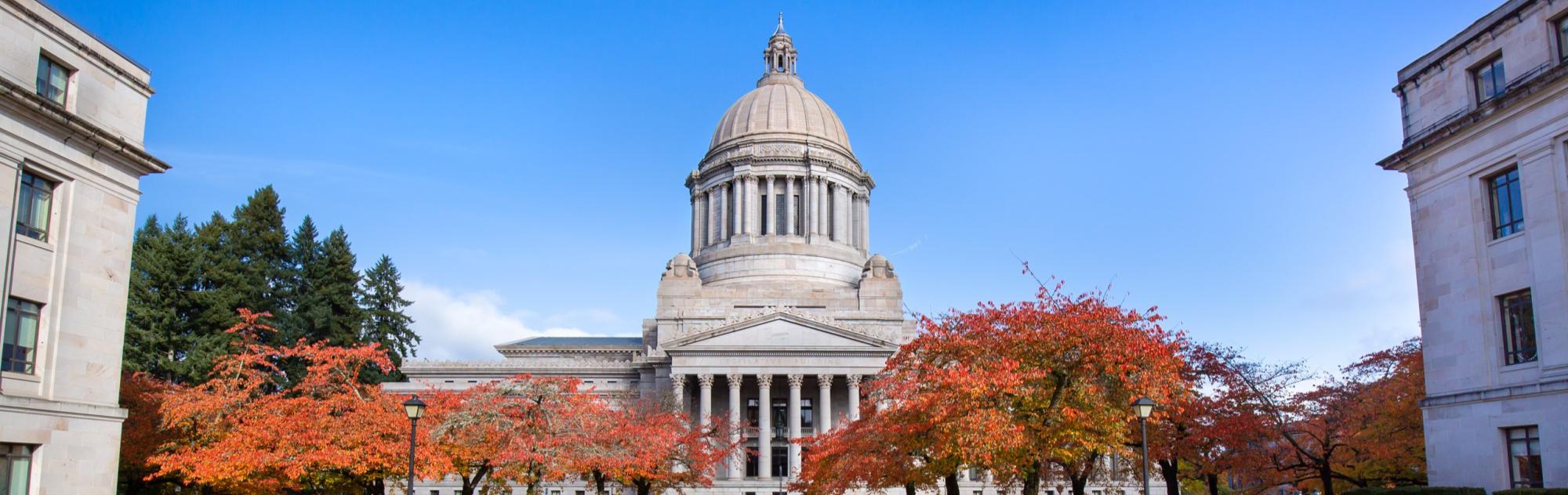 The Legislative Building, also known as the Capitol Building, with orange and red fall trees in the foreground and a blue sky in the background.