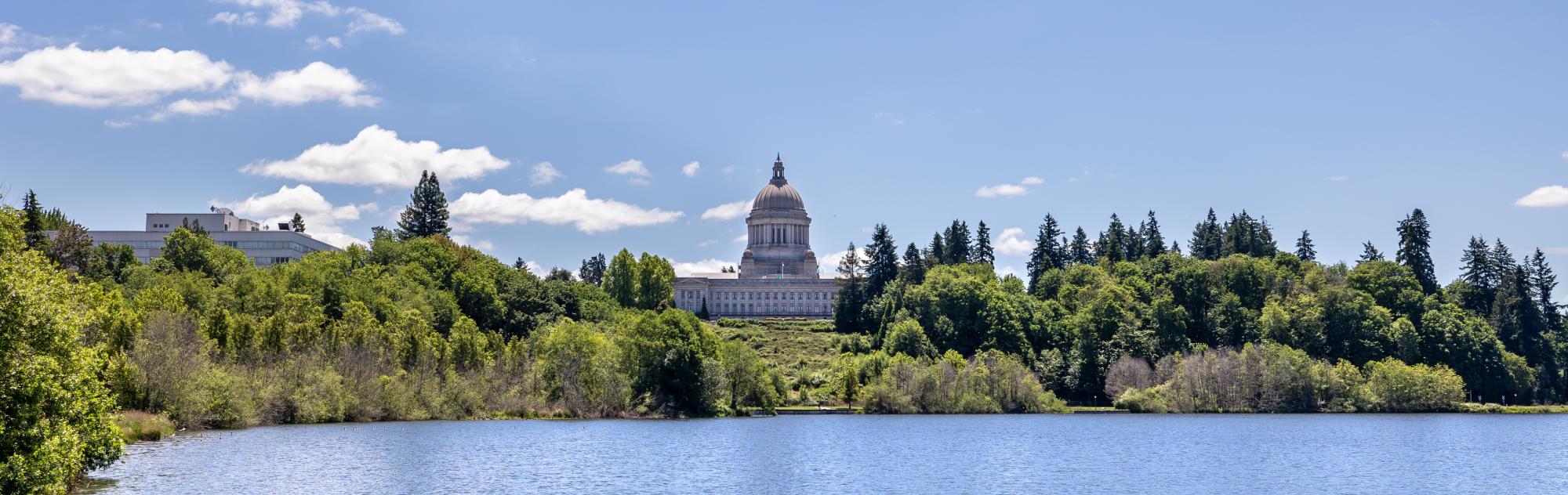 An outdoor scene showing a blue lake surrounded by large green trees on a sunny day. The bright white Legislative Building dome stands prominently in the treeline.