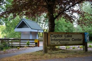 A nature scene with a sign that says "Capitol Lake Interpretive Center, State of Washington" in the foreground and a cement pathway, a wooden structure, and trees in the background.