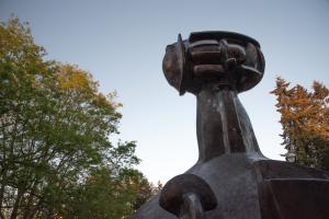 The piece at the top of the Shaman Statue has a strong, dark silhouette against the light blue sky and the vibrant trees around it.