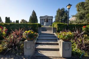 A nature scene featuring a set of the Sunken Garden's steps and pillars, flowers and bushes, and the Winged Victory Monument in the background.