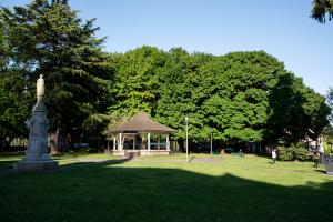 A nature scene featuring an open lawn, a gazebo, a granite statue, people sitting on park benches, and large trees in the background.