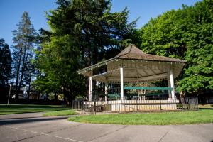 A nature scene with a cement pathway leading to a wooden gazebo sitting in a patch of grass, with large trees in the background.