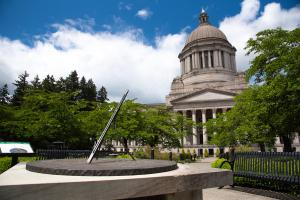 An outdoor scene with the Territorial Sundial in the foreground, a stone pathway lined with metal park benches and cherry blossom trees, and the Legislative Building in the background.