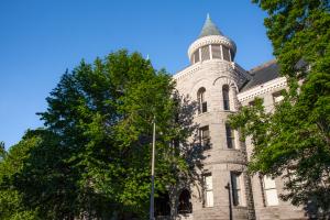 The back of the Old Capitol Building, which has two towers with teal cone-shaped roofs, similar to the front side of the building. There are three large trees with green leaves and one streetlight outside of the back of the building. 