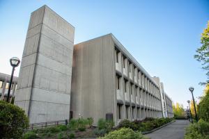 A view of the back corner of the Department of Transportation Building from the southeast. A large, rectangular concrete pillar stands tall above the rest of the building, and various green trees, shrubs, and other plants line the pathway that leads to the back entrance of the building.