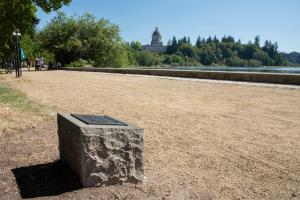 The Women Veterans marker, which sits on the side of a gravel pathway at Capitol Lake. The Legislative Building dome is in the background, surrounded by trees.