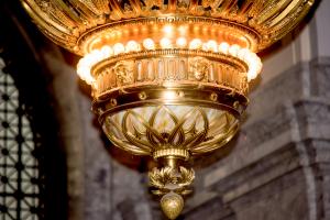A close-up of the bottom portion of the large chandelier that hangs in the Legislative Building Rotunda. This section features small faces and flowers, white and gold marble, and a single row of small light bulbs.