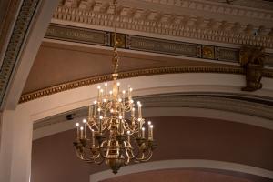 A polished bronze chandelier with lights that resemble candles. The chandelier hangs in the corner of a Legislative chamber room.
