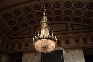 A polished bronze chandelier with thousands of glass beads on it hanging from a patterned ceiling. 