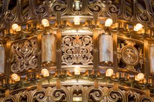 A close-up of the middle portion of the large chandelier that hangs in the Legislative Building Rotunda. This section features mythological creatures, an eagle, floral elements, and two rows of small light bulbs.