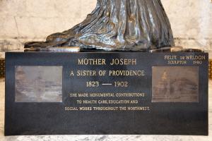 A rectangular, black granite platform inscribed with information about Mother Joseph. Two etched pictures of old buildings are on metal plaques on the sides of the inscribed text.