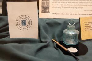 A piece of paper with a postage stamp and a drawn seal design on it, an old ink bottle, an old ink pen, and a silver dollar inside a glass display case..