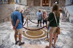 A tour guide and tourists standing around a large bronze Washington State Seal roped off in the middle of a marble floor.