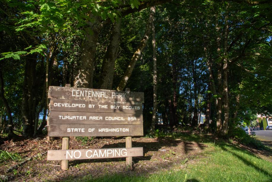 A nature scene with a sign that says "Centennial Park, Developed by the Boy Scouts, Tumwater Area Council BSA, State of Washington, No Camping" in the foreground and lush, leafy trees in the background.