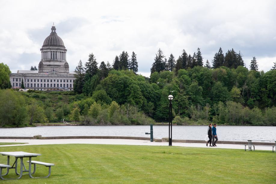 A nature scene with an open lawn with picnic tables in the foreground, people walking on a gravel pathway in front of Capitol Lake, and trees and the Legislative Building in the background.