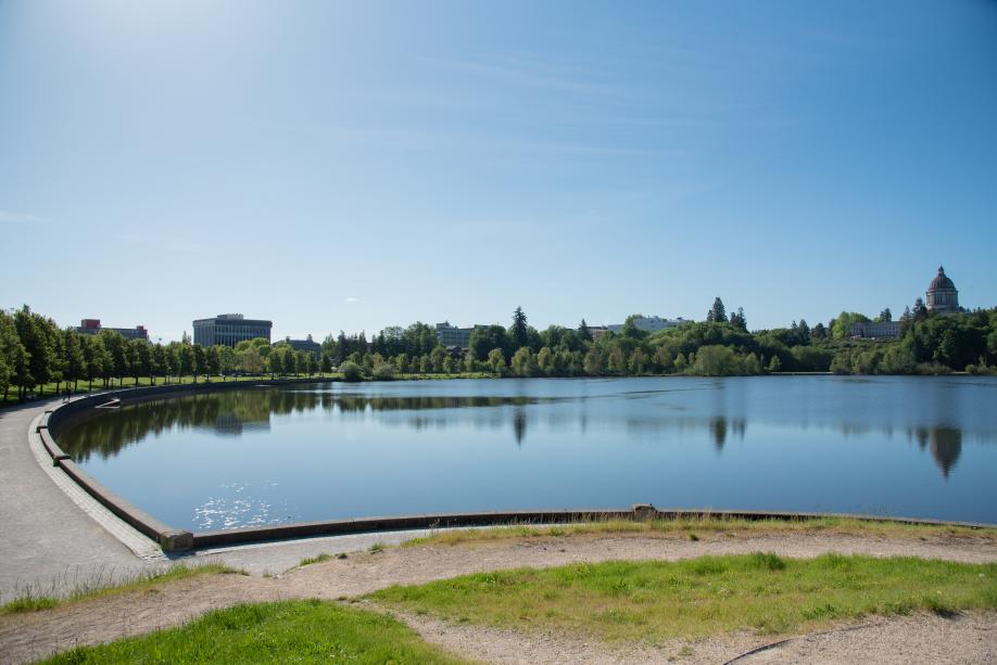 A nature scene predominantly featuring Capitol Lake, with a gravel pathway lined with trees and the Legislative Building in the background.