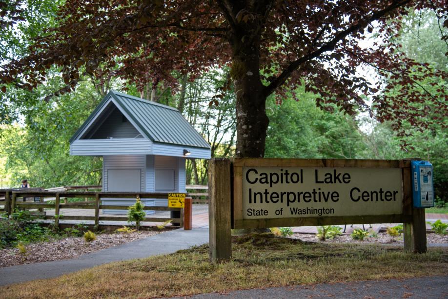 A nature scene with a sign that says "Capitol Lake Interpretive Center, State of Washington" in the foreground and a cement pathway, a wooden structure, and trees in the background.