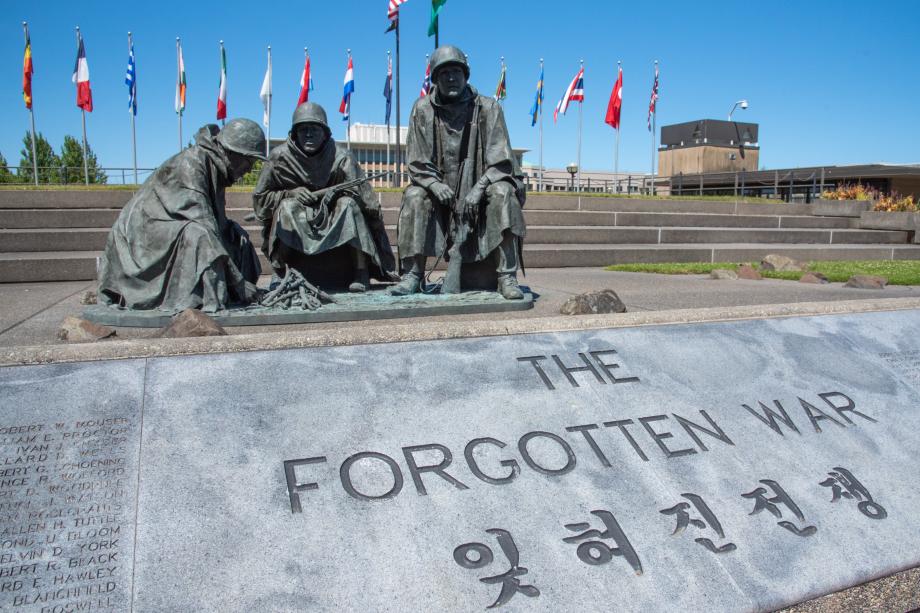 A close-up of a stone surface inscribed with "The Forgotten War" in front of a bronze statue depicting three soldiers sitting by a campfire, with the flags of various countries behind them.