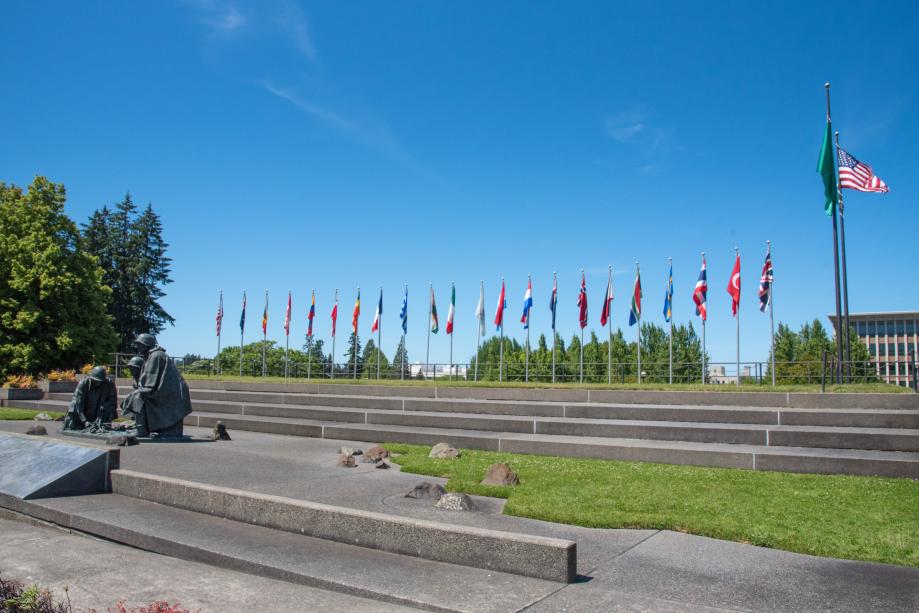 An outdoor scene with a stone pathway leading to the Korean War Memorial statue and steps leading to multiple flagpoles featuring the flags of the different countries that participated in the Korean War.