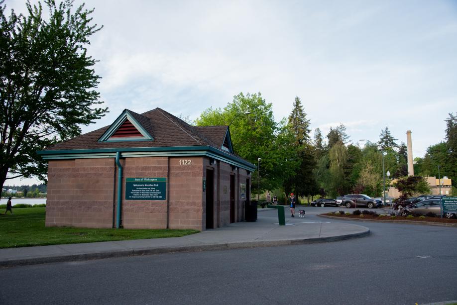 An outdoor scene featuring a brick building (the Marathon Park restrooms), a woman walking her dog, various trees, and a full parking lot in the background.