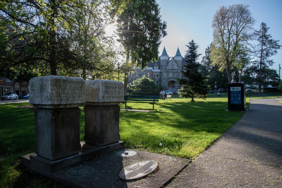 A nature scene featuring two small, marble water fountains set off a cement pathway that leads through an open lawn decorated with park benches, with the Old Capitol Building in the background.