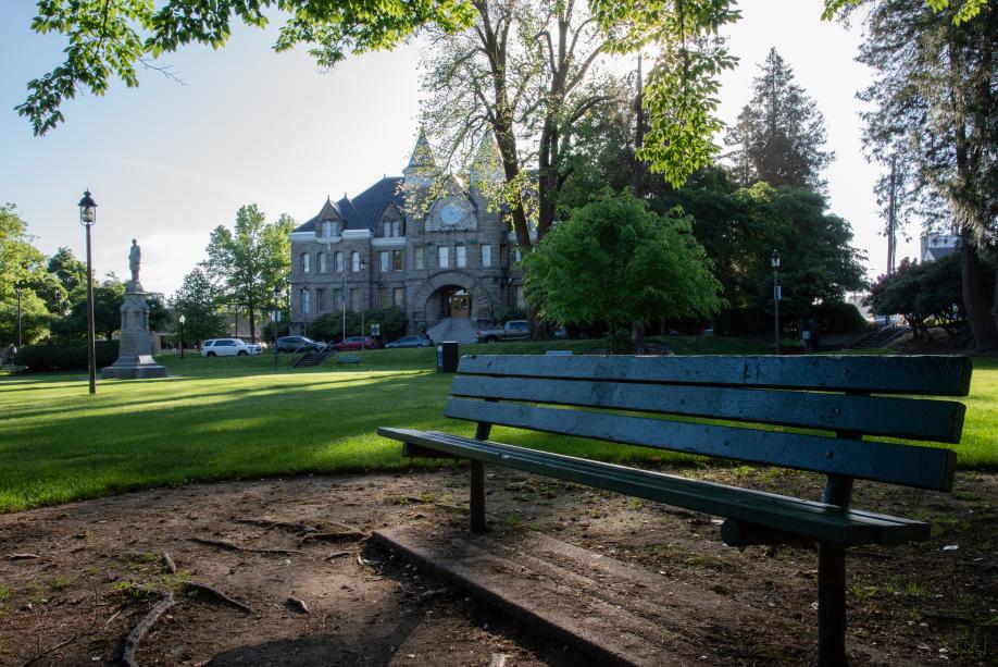 A nature scene with a wooden park bench shaded by a large tree in the foreground and an open lawn, trees, a granite statue, and the Old Capitol Building in the background.