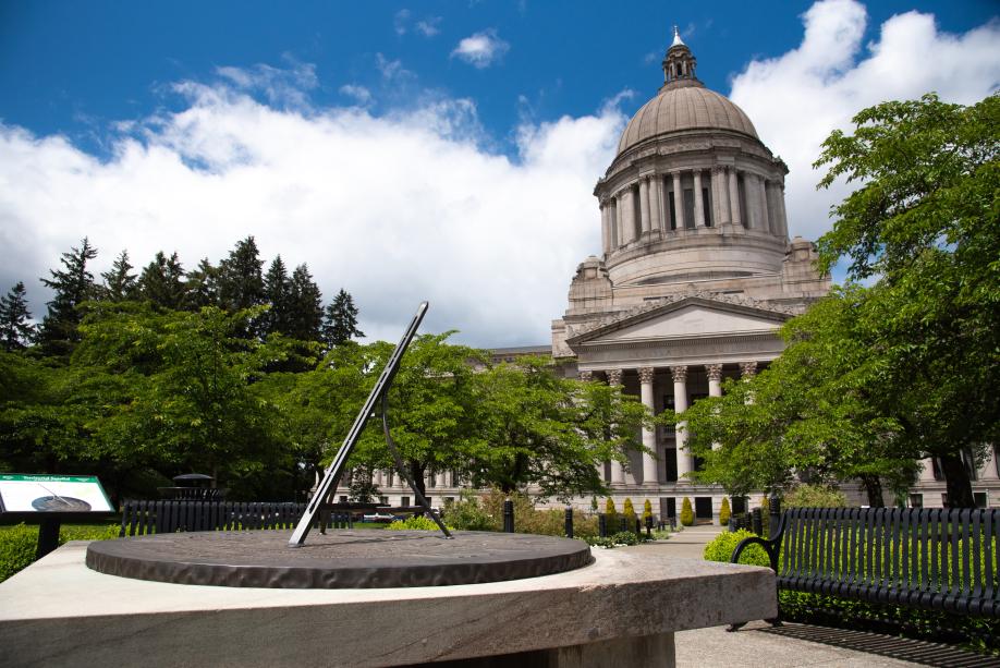 An outdoor scene with the Territorial Sundial in the foreground, a stone pathway lined with metal park benches and cherry blossom trees, and the Legislative Building in the background.