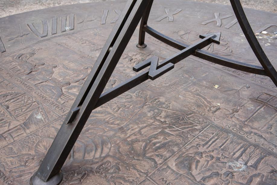 A close-up of the Territorial Sundial's bronze rods, one of which is decorated with the letter "N" to signify North, and the brass platform with images and text engraved into it.