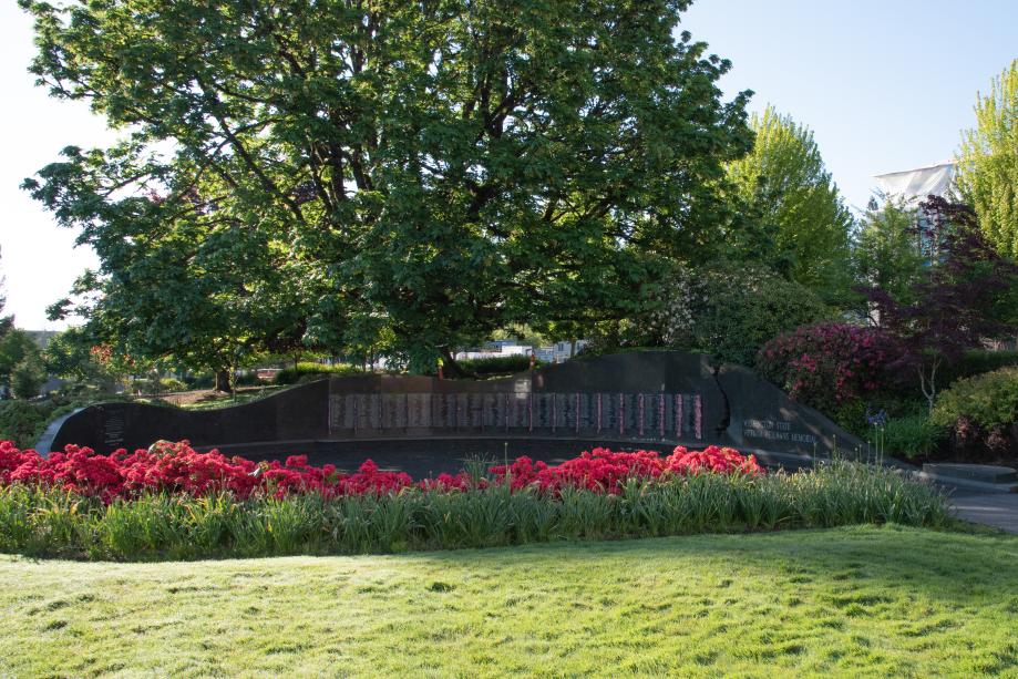 A nature scene with red flowers in the foreground surrounding the Vietnam Veterans Memorial with a large green tree and floral bushes in the background.