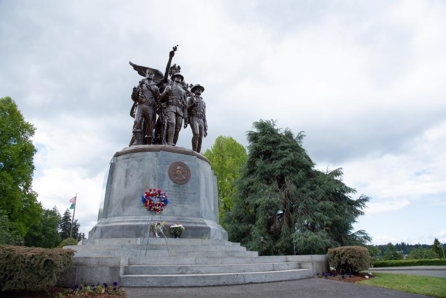 An outdoor scene with flowers placed on the steps of the Winged Victory Monument with trees and an American flag in the background.