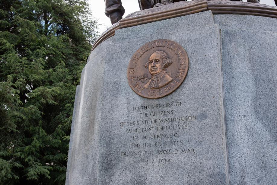 A close-up of the bronze Washington State Seal set into the granite base of the Winged Victory Monument with trees in the background.