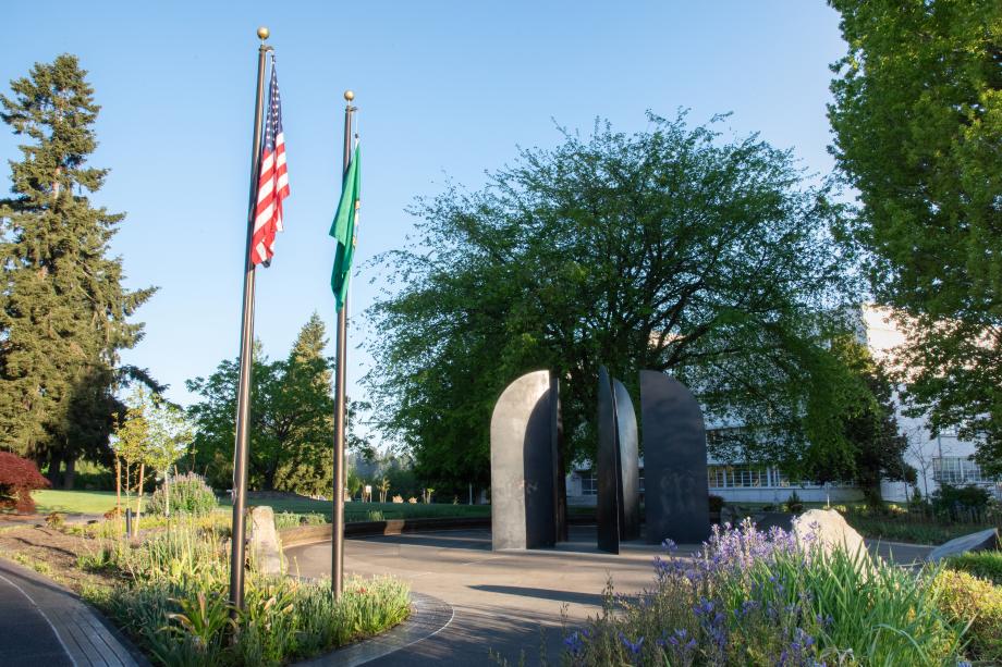 A nature scene with the American flag and Washington State flag in the foreground set off a stone pathway leading to the World War II Memorial's bronze blades, surrounded by flowers and large trees.