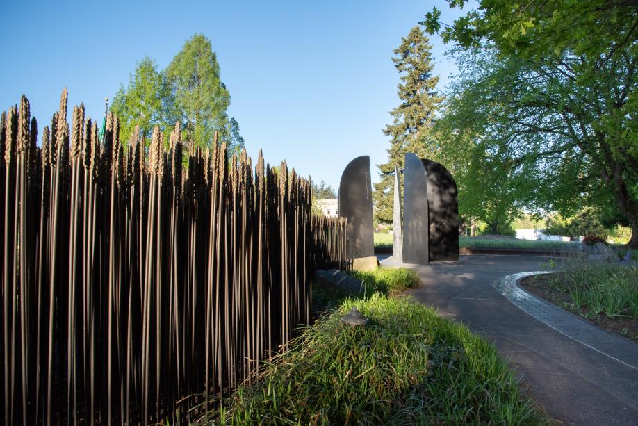 A nature scene with a bronze wheat patch in the foreground next to a stone pathway that leads to the World War II Memorial's bronze blades, surrounded by large green trees.