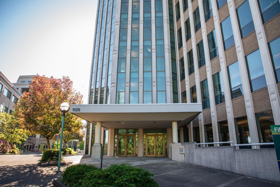 The south entrance to the Highways-Licenses Building, with the numbers "1125" above the doors. A small concrete plaza with green shrubs and a reddish-orange trees sits between this entrance and the Natural Resources Building entrance.