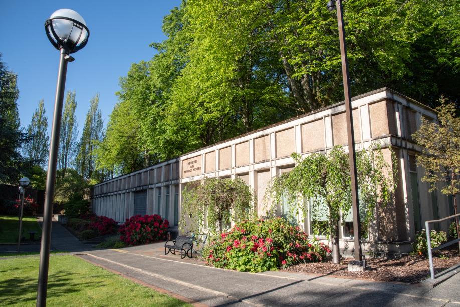 A view of the front of the State Archives Building from the northeast. A stone pathway in front of the building is lined with various plants and light posts, and large trees with green leaves are growing on the roof of the State Archives building.