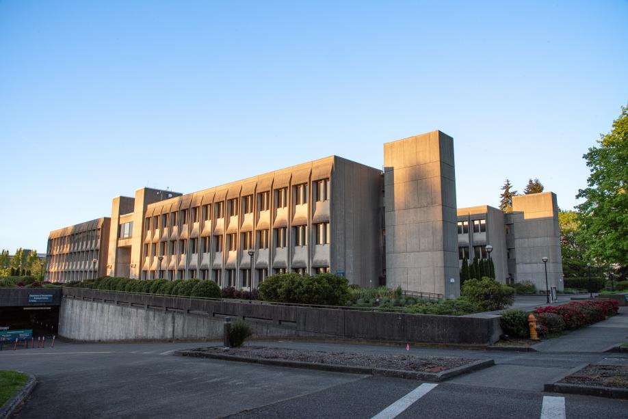 A street view of the Department of Transportation Building from the southwest. Large, rectangular concrete pillars at the ends of the building stand tall above the rest of the structure, with trees and other plants around it.