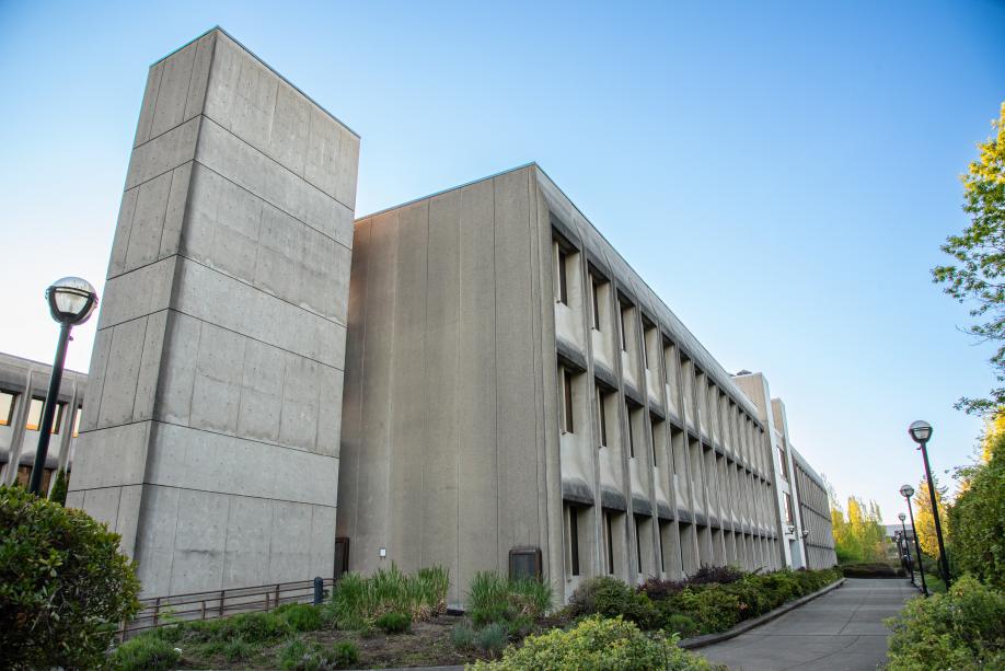 A view of the back corner of the Department of Transportation Building from the southeast. A large, rectangular concrete pillar stands tall above the rest of the building, and various green trees, shrubs, and other plants line the pathway that leads to the back entrance of the building.