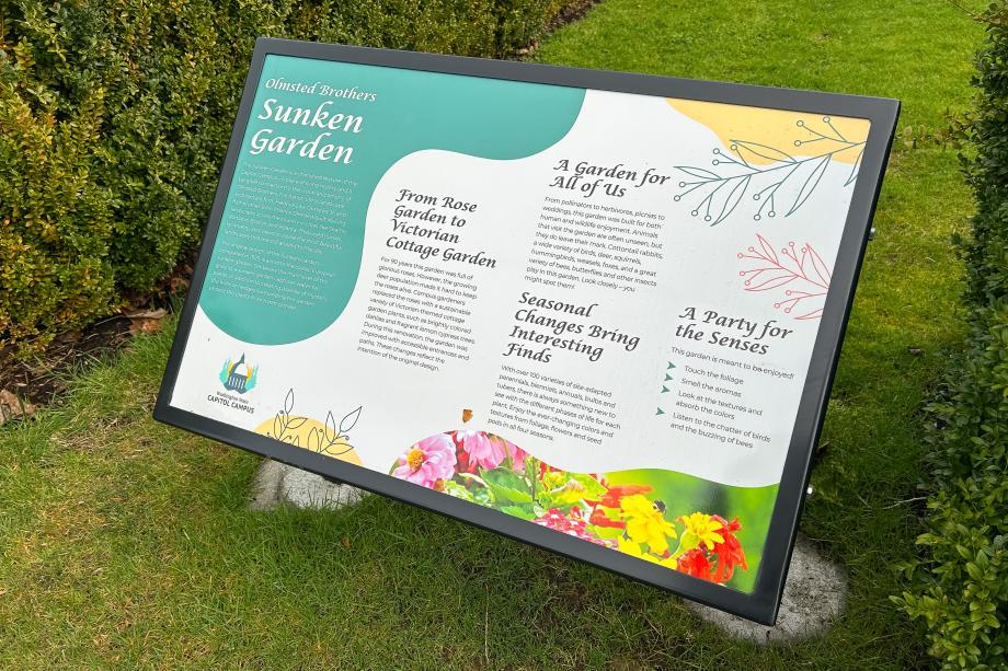 An overhead view of a large, colorful sign that explains the Sunken Garden. The sign stands in the grass between two bushes on the south edge of the Sunken Garden.