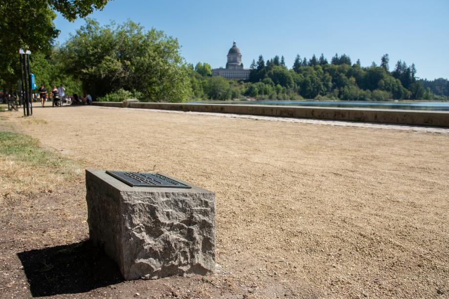 The Women Veterans marker, which sits on the side of a gravel pathway at Capitol Lake. The Legislative Building dome is in the background, surrounded by trees.