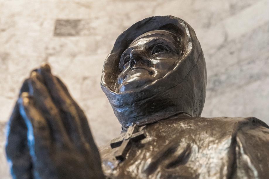 A close-up of the Mother Joseph statue, focusing on her face and hands. She wears a cross necklace around her neck, and she's looking up toward the ceiling.