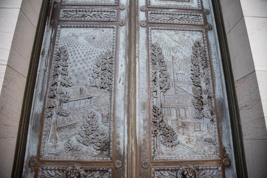 A pair of bronze doors with carved images on them. The left door shows a house surrounded by hills and trees, and the right door shows an old capitol building also surrounded by hills and trees.