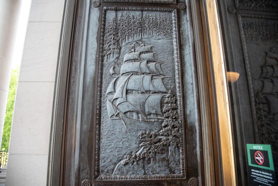 A bronze door with a carved image of a large ship sailing through a body of water surrounded by evergreen trees.