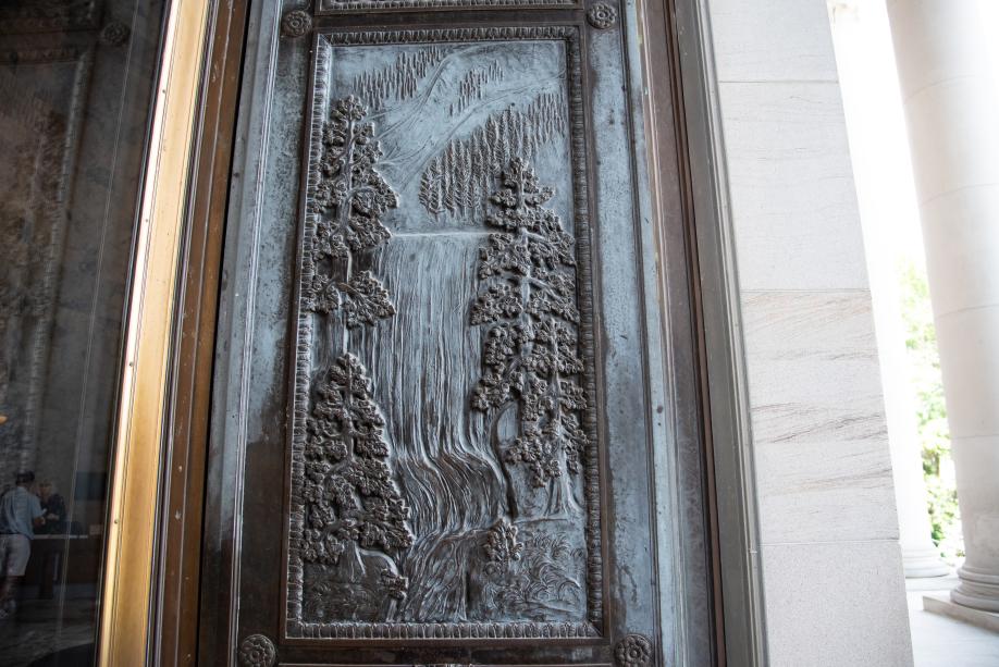A bronze door with a carved image of a waterfall in a forest full of evergreen trees.