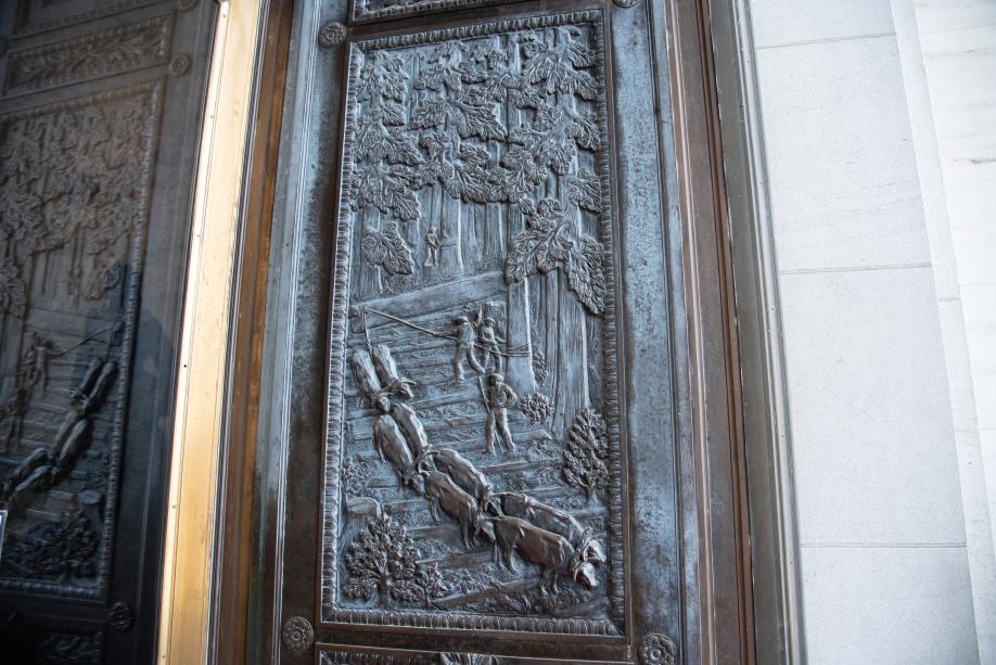 A bronze door with a carved image of a logging crew cutting down large trees while oxen pull the logs.