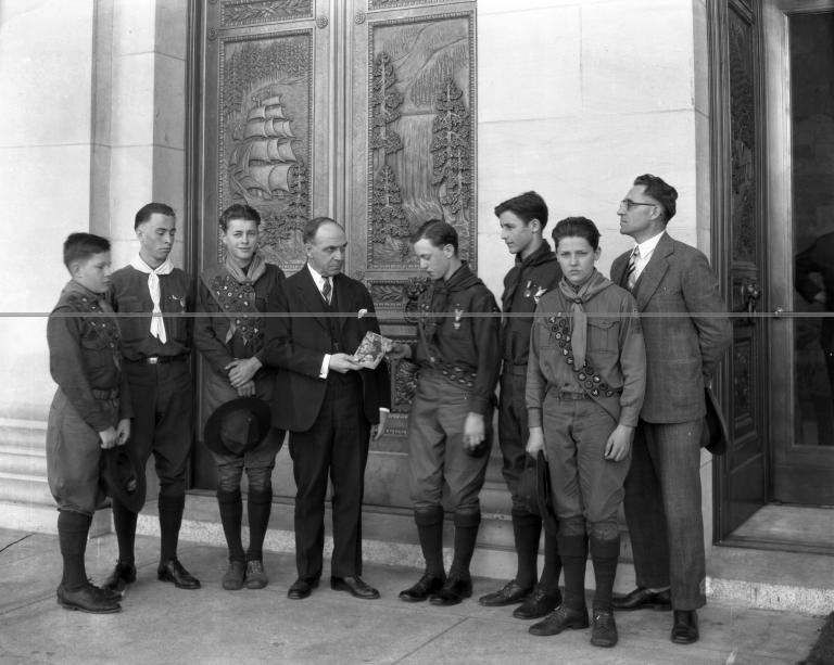 A black and white photo of a governor standing with six boy scouts and another government official in front of the large bronze doors of the Legislative Building.